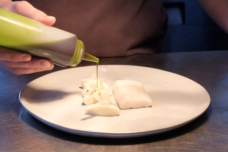 Close up of male chefs hands pouring green sauce from a clear plastic bottle onto a white plate with white fish on it, the plate is on a silver metal kitchen top behind is the part of the grey apron the chef is wearing in a fine dining restaurant.の写真素材