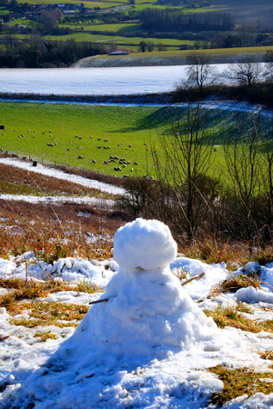 A small snowman with sticks as arms sits on the edge of snow covered rolling hills of the South Downs national park, East Sussex, UK, the village of Glynde is in the background the sun is shining on the multicoloured fieldsの写真素材