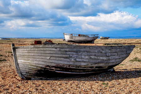 an old wooden fishing boat sitting on a pebble beach, in the back ground on the beach are two more old wooden fishing boats, the pebbel beach is flat and big with a big cloudy blue sky, Dungeness, Kent, England, United Kingdomの写真素材