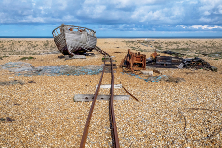 Old rusty train track on a big flat pebble beach leading to an old wooden fishing boat that is sitting on the beach, scattered across the beach are old fishing nets, winching equipment and wood, in the background there is a thin line of blue sea and above is a blue cloudy sky.の写真素材