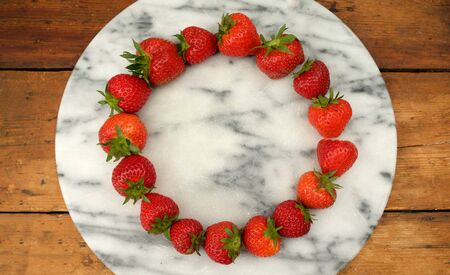sixteen red ripe strawberries positioned in a circular shape on a marble circular board in the middle of the circle of strawberries is a large strawberry on a rustic wooden table top, still life with lots of copy spaceの写真素材