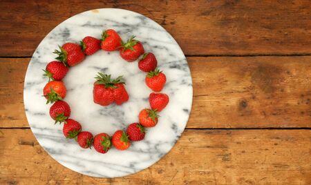 sixteen red ripe strawberries positioned in a circular shape on a marble circular board in the middle of the circle of strawberries is a large strawberry on a rustic wooden table top, still life with lots of copy spaceの写真素材
