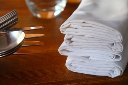A restaurant table on the left are knives, forks and spoons stacked ontop of each other on the right four white linen napkins, wooden table, warm colours.の写真素材