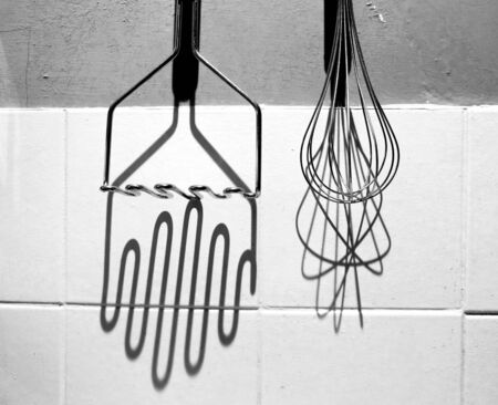 Black and white photograph of a silver metal potato masher and hand whisk hanging up on a kitchen...