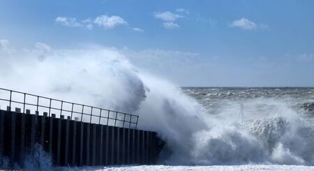 Dramatic stormy sea breaking against a harbour wall, spray and waves high in the air, rough sea and a blue sky in the distance, Seaford, East sussex, England, UK,の写真素材