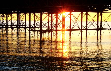 The metal support posts of Brighton pier rising out of ocean, the sun is setting under the pier forming a beautiful star burst effect of orange light, the metal post form graphical shapes glowing orange on a calm seaの写真素材