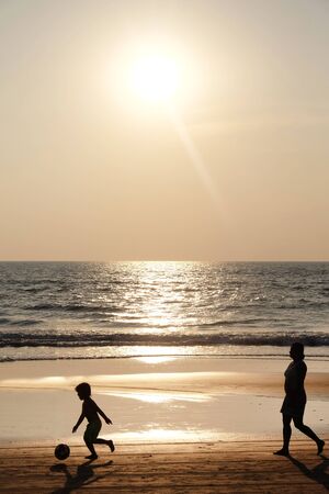 Silhouette of an unrecognizable small child holding a football, followed by his mom, they are both walking along an empty golden sandy beach at sunset, a calm sea and clear golden sun lit sky の写真素材