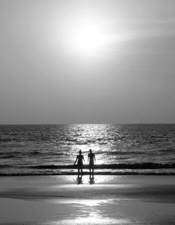 Black and White Silhouette of an unrecognizable young couple standing together on an empty golden sandy beach looking at the sunset, a calm sea and clear golden sun lit sky の写真素材