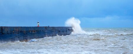 A harbor wall with a rough stormy sea crashing against the wall causing the sea to be blurred and in motion, behind is a red and white lighthouse, waves are crashing over the wall, there is a blue sky behindの写真素材