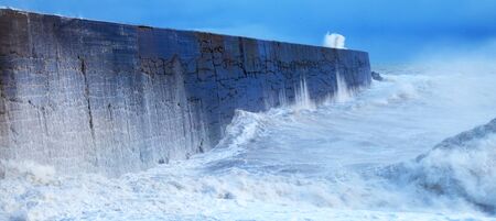 A harbor wall with a rough stormy sea crashing against the wall causing the sea to be blurred and in motion, waves are crashing over the wall, there is a blue sky behindの写真素材
