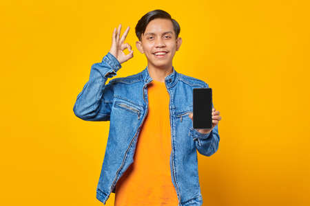 portrait of young asian man holding smartphone and showing ok sign over yellow backgroundの写真素材