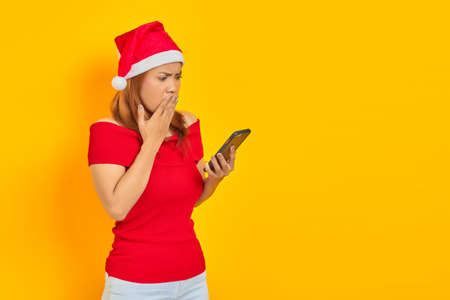 Surprised young Asian woman in Santa Claus hat looking at mobile phone with open mouth isolated over...