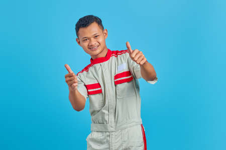 Portrait of excited young Asian mechanic showing thumb up gesture isolated on blue backgroundの写真素材