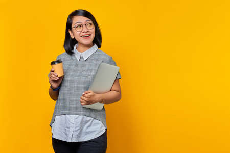Portrait of smiling young Asian woman wearing glasses holding laptop and coffee looking sideways...
