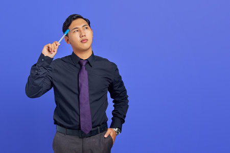 Serious young handsome businessman thinking about something and holding pen on purple backgroundの写真素材