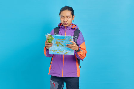 Portrait of Asian young man wearing jacket and carrying bag looking serious reading map isolated on...