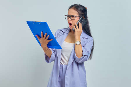 Portrait of surprised young Asian woman talking on mobile and looking at task list in documents...