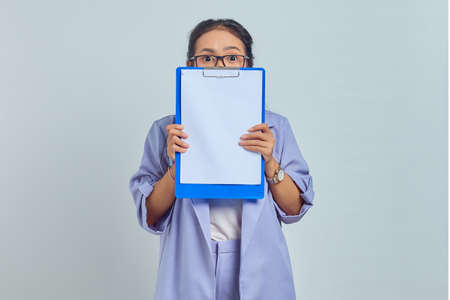 Portrait of scared young Asian business woman covering face with folder isolated on purple...