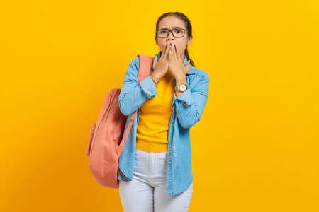 Surprised young Asian student in denim clothes with backpack covering mouth with hands isolated on yellow background. Education in high school university college conceptの写真素材