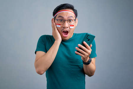 Shocked young Asian man in casual t-shirt  using mobile phone, reacting to new app isolated on grey...