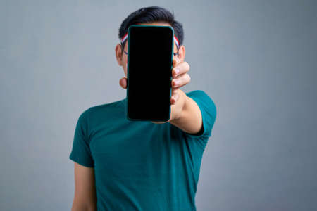 Handsome young Asian man in casual t-shirt showing mobile phone with blank screen, recommending mobile app isolated on grey background. indonesian independence day celebration conceptの写真素材
