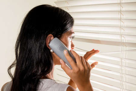 a young woman watching something through the blinds of her windowの写真素材