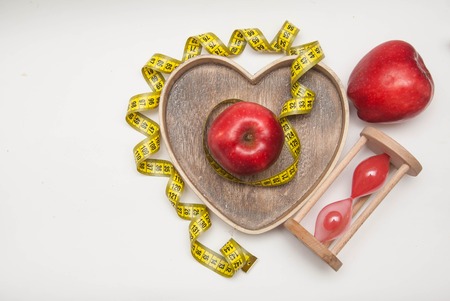 Diet and Healthy Food. Glass Clock and Red ripe Apple in Heart Shape Wooden Box, tied with Cyan Measuring Tape, isolated on white background with copy spaceの写真素材