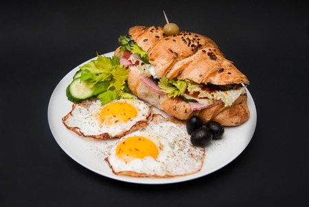 Fried Eggs with Croissant Sandwich and Fresh Vegetables over Black Background.の写真素材