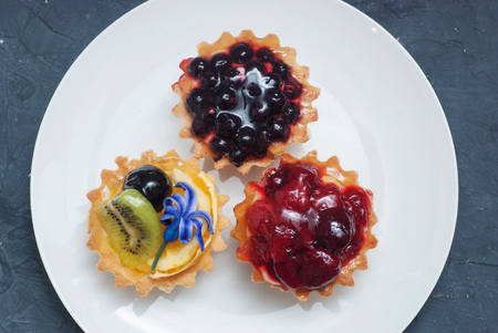Three Tarttlets with Cream and Fruits, Berries on White Round Plate over Dark Background.の写真素材