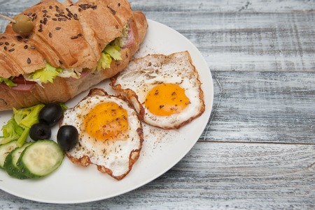 White Plate with Fried Eggs and Croissant Sandwich with Cucumber, Tomatoe and Olives. Breakfast. Gray Wooden Background.の写真素材