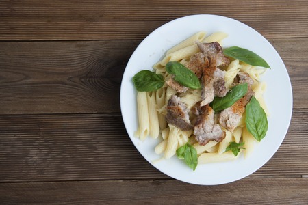 Cooked penne pasta isolated in white round plate, with basil leaves and pork meat, over wooden table, decorated with basil leaves.の写真素材