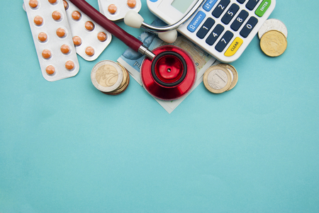Red stethoscope on a pile of coins, pills over blue background.の写真素材