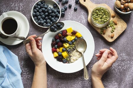 Healthy breakfast with oatmeal porridge, fruits and nuts. Blueberry, mango, pumpkin seeds, nuts, coffee cup. Top view. Female hands.の写真素材