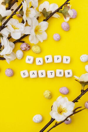 Happy Easter. Cubes with text on yellow background. Spring brunches with white flowers and candy, chocolate easter eggs.の写真素材