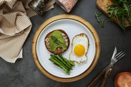 Avocado toasted sandwich and a fried egg on a white plate with asparagus. Healthy food or breakfast. Dark board.の写真素材