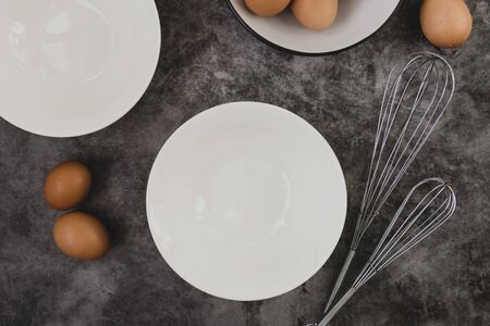 Dark baking background with eggs, whisk, eggshell and white plates. Copy space, top view for text.