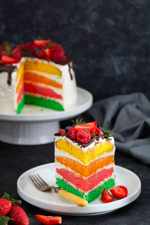 Rainbow slice of cake with fresh berries on dark background.