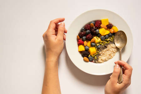 Healthy breakfast with oatmeal porridge, fruits and nuts. Blueberry, mango, pumpkin seeds, nuts, coffee cup. Top view. Top view white background. Female hands holding the plate.の写真素材