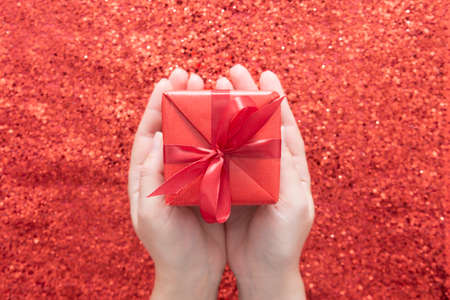 Female hands holding gift box over red glitter sparkling background, copy space. Valentine's Day, Christmas.の写真素材