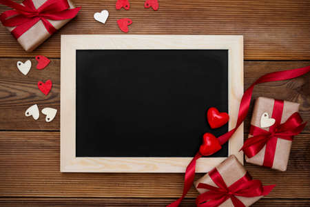 Gift boxes with red ribbon and empty chalkboard on wooden table.