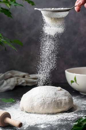 Cooking sourdough homemade bread. Sieving flour over raw bread dough on table, dark background,...