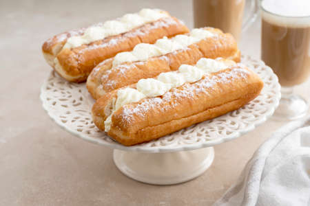 Whipped cream eclairs and coffee cups, on warm neutral background. Breakfast food