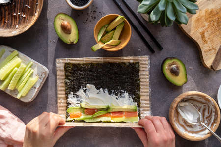 Cooking Sushi rolls at home with salmon, avocado and cucumber. Female hand preparing avocado sushi, top view. Step by step.の写真素材