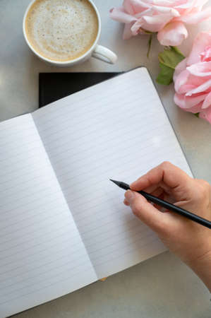 Open note book and female hands holding a pencil. Empty page mockup, cup of coffee, pink roses...
