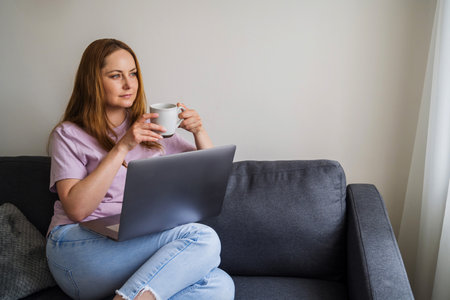 Woman working online, sitting on a sofa, drinking a coffee in living room and using laptopの写真素材