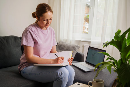 Woman taking notes in a notebook, sitting on a sofa in living room and using laptopの写真素材
