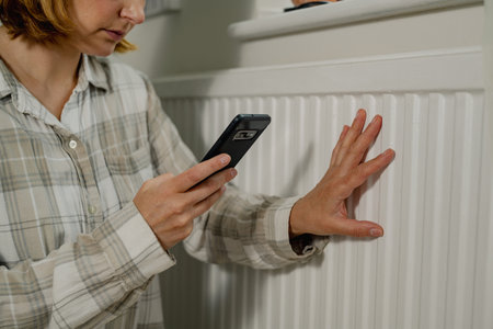 Woman holding smart phone next to heating thermostat, increasing heating costs