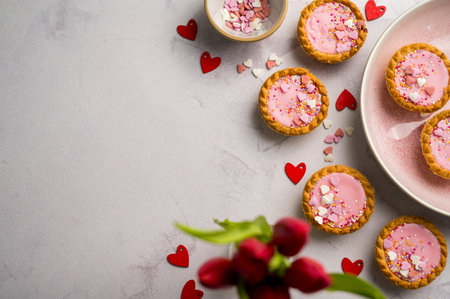 Valentine's day background. Pink mini tarts with heart shaped sprinkles, red hearts and red tulips. Top view, copy spaceの写真素材