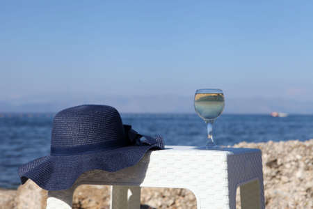 A glass of white wine and a blue hat behind lagoon sea. Summer vacation concept. glass of wine and blue hat near the seaの写真素材