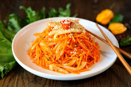 Carrot Korean squid meat salad in a plate with chopsticks on wooden oak tableand green salad leafs, fresh yellow pepper. Spicy Korean-style carrot salad on a dark background. Koryo-saram cuisine.の写真素材